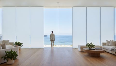 Oceanfront Hermosa Beach great room with a floor-to-ceiling switchable smart glass wall framing The Strand and the pier — half the panels crystal clear to the Pacific, half switched to flat, even, milky-white frosted privacy mode