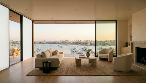 Luxury Orange County coastal living room with a floor-to-ceiling switchable smart glass wall opening onto a Newport Beach harbor view at golden hour — several glass panels switched to frosted privacy mode