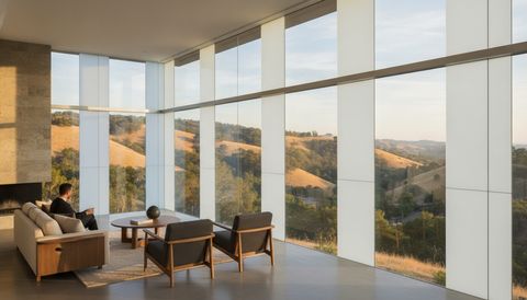 Modern Conejo Valley hillside living room with a wall of switchable smart glass framing the oak-covered hills — half the panels crystal clear, half switched to flat, even, milky-white frosted privacy mode
