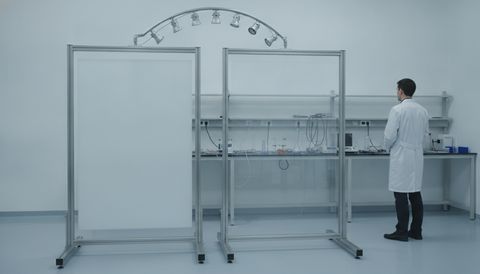 Illustration of two switchable smart film sample panels racked side by side in a bright testing lab, one frosted an even flat milky white and one clear, beneath an angled arc-lamp rig