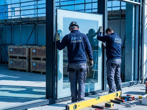 Installers seen from behind fitting a large clear smart glass panel into a black aluminum frame on a jobsite, with crated smart glass panels ready in the background