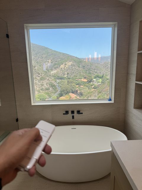 Hand holding a Smart View remote in a Laguna Beach bathroom, the picture window clear with the canyon hillside beyond