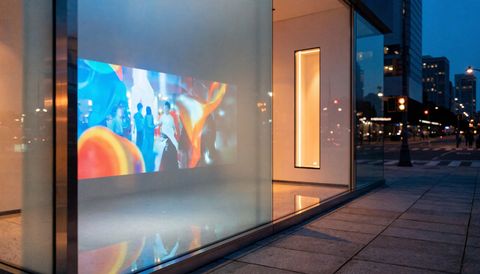 Illustration: frosted smart glass storefront at dusk carrying a bright rear-projected ad while the space glows behind it